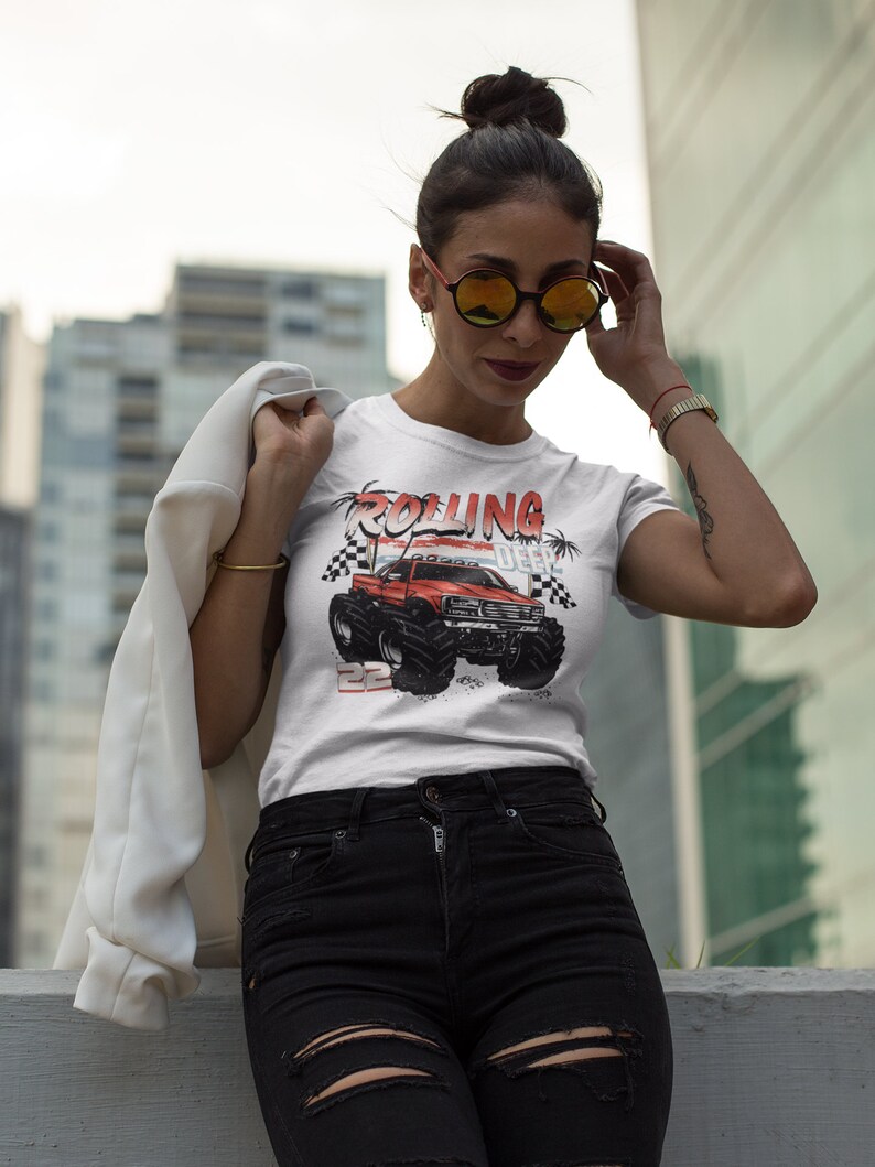 May include: White t-shirt with a red monster truck graphic and the text "Rolling Deep". The truck has black wheels and a red body. The t-shirt is worn by a person with black hair and sunglasses.