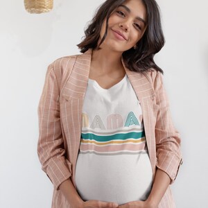 May include: A pregnant person wearing a white t-shirt with the word "MAMA" in colourful, retro-style lettering and a pink striped blazer. The person is smiling and holding their belly.