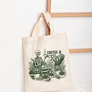 May include: A white tote bag with a green illustration of a skeleton sitting in a chair with a cup of coffee and surrounded by plants. The text "Coffee & Plants" is written in green above the skeleton.