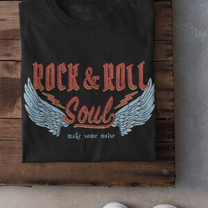 Puede incluir: Camiseta negra con un dise&ntilde;o gr&aacute;fico de estilo vintage que presenta el texto "Rock & Roll Soul" con alas y rayos. El texto "make some noise" est&aacute; debajo del gr&aacute;fico.