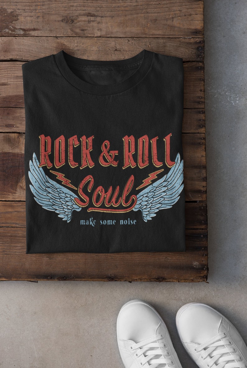 Puede incluir: Camiseta negra con un dise&ntilde;o gr&aacute;fico de estilo vintage que presenta el texto "Rock & Roll Soul" con alas y rayos. El texto "make some noise" est&aacute; debajo del gr&aacute;fico.