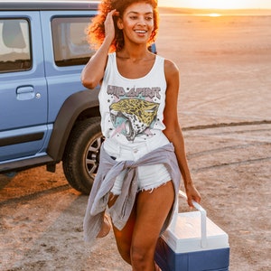 May include: A woman wearing a white tank top with a graphic of a jaguar and the text "Wild Spirit" in pink and white. She is wearing white denim shorts and a grey shirt tied around her waist. She is holding a blue and white cooler.