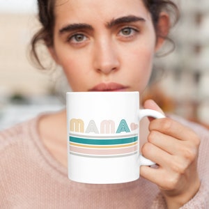 May include: White ceramic mug with the word "MAMA" in teal, grey, and pink lettering with a small pink heart. The mug has a teal, grey, and pink striped design.