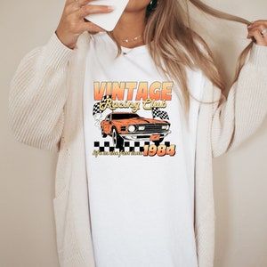 a woman wearing a t shirt with the vintage racing club design. the retro design features a orange muscle car with checkered racing flags.  the words vintage racing club above and the words life in the fast lane 1984 below.
