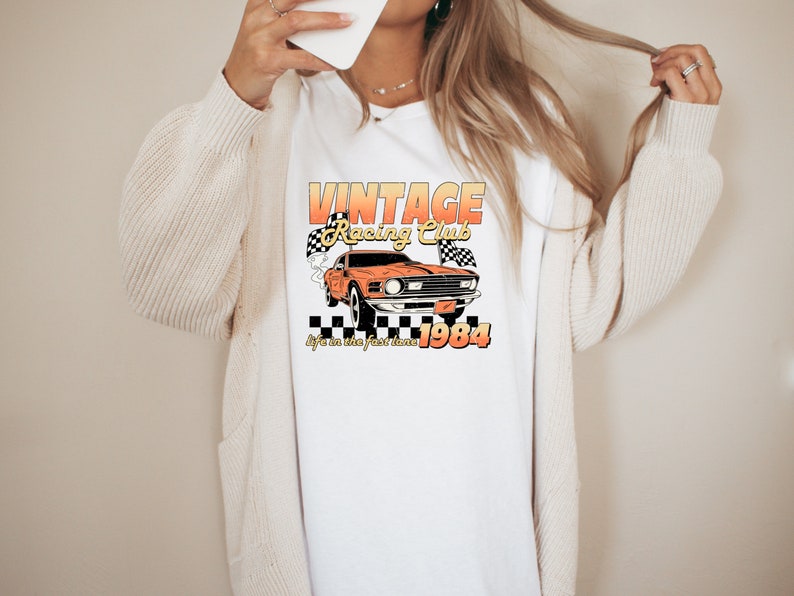 a woman wearing a t shirt with the vintage racing club design. the retro design features a orange muscle car with checkered racing flags.  the words vintage racing club above and the words life in the fast lane 1984 below.