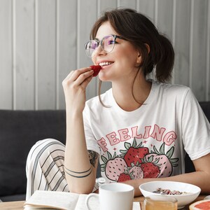 Puede incluir: Una mujer con gafas y una camiseta blanca con el texto "Feeling" y un gr&aacute;fico de fresas est&aacute; comiendo una fresa. Est&aacute; sentada en una mesa con un libro, una taza de caf&eacute;, un taz&oacute;n de cereal y un pomelo.