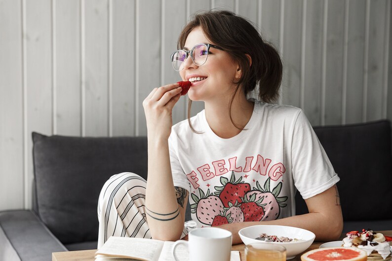 Puede incluir: Una mujer con gafas y una camiseta blanca con el texto "Feeling" y un gr&aacute;fico de fresas est&aacute; comiendo una fresa. Est&aacute; sentada en una mesa con un libro, una taza de caf&eacute;, un taz&oacute;n de cereal y un pomelo.