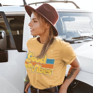 May include: A woman wearing a yellow t-shirt with the text "Good Vibes" in a retro font and colorful stripes. She is wearing a brown hat and blue jeans.