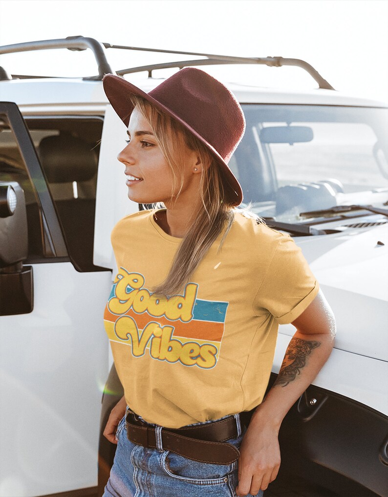 May include: A woman wearing a yellow t-shirt with the text "Good Vibes" in a retro font and colorful stripes. She is wearing a brown hat and blue jeans.