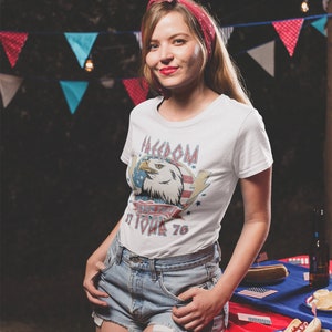 May include: A woman wearing a white t-shirt with a vintage American eagle graphic and the text "Freedom Isn't Free It Took 76". She is wearing denim shorts and a red bandana in her hair. She is standing in front of a red, white, and blue tablecloth with hot dogs on it.