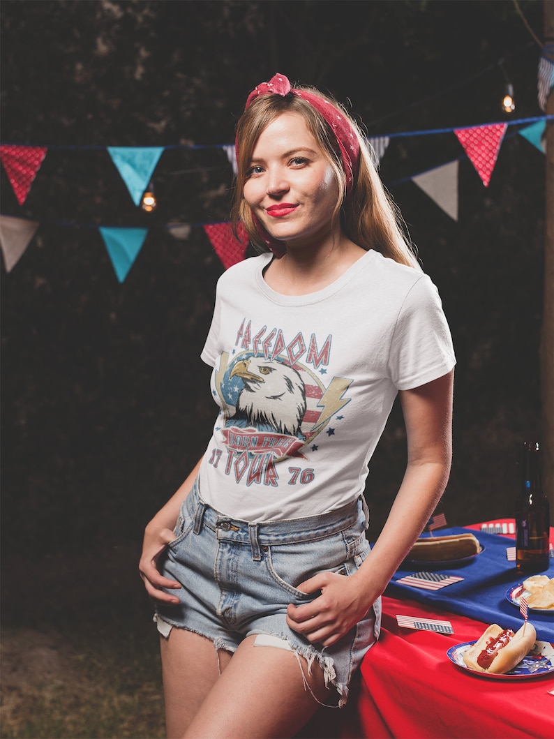 May include: A woman wearing a white t-shirt with a vintage American eagle graphic and the text "Freedom Isn't Free It Took 76". She is wearing denim shorts and a red bandana in her hair. She is standing in front of a red, white, and blue tablecloth with hot dogs on it.