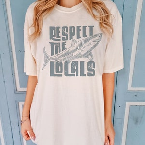 a woman wearing an oversize t shirt.  the shirt features a graphic of a shark and the words "respect the locals" in a retro text.  she is standing in front of a blue door.