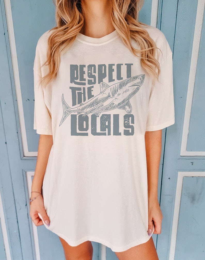 a woman wearing an oversize t shirt.  the shirt features a graphic of a shark and the words "respect the locals" in a retro text.  she is standing in front of a blue door.
