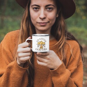 Pu&ograve; includere: Una tazza da campeggio in smalto bianco con manico nero e il testo "Life is better by a campfire" in lettere marroni e gialle. La tazza &egrave; decorata con una scena di fal&ograve; con montagne e alberi.
