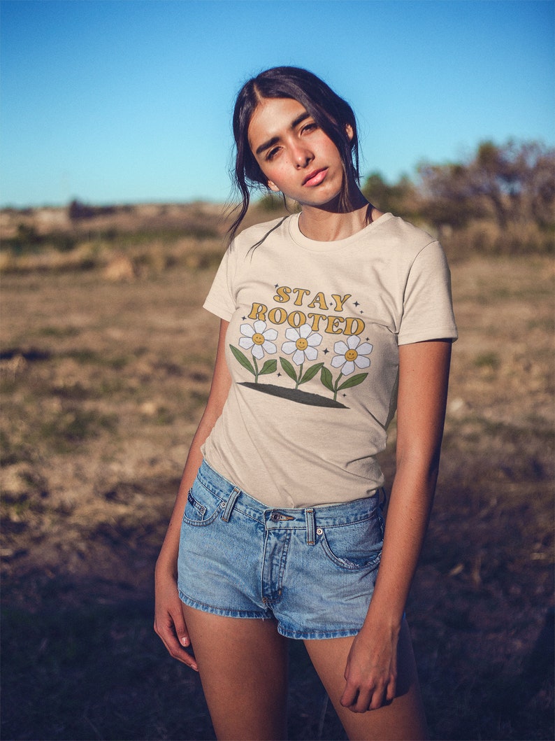 May include: A woman wearing a beige t-shirt with the text "STAY ROOTED" in yellow and three white flowers with yellow centers. She is wearing blue denim shorts.