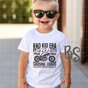 以下が含まれることがあります： 「Rad Kid Era Causing Chaos Life in the Fast Lane」というテキストと、黒いオートバイのグラフィックが描かれた白いTシャツです。