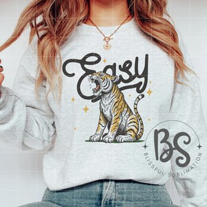 May include: A gray sweatshirt with the text "Easy" in a stylized font, featuring a roaring tiger with yellow and orange stripes. The text "BS" is below the tiger, with the words "Blissful Sublimation" written around it.