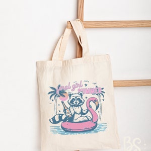May include: A white tote bag with a pink flamingo pool float and a raccoon holding a bottle of beer. The text "feral girl summer" is printed on the bag.
