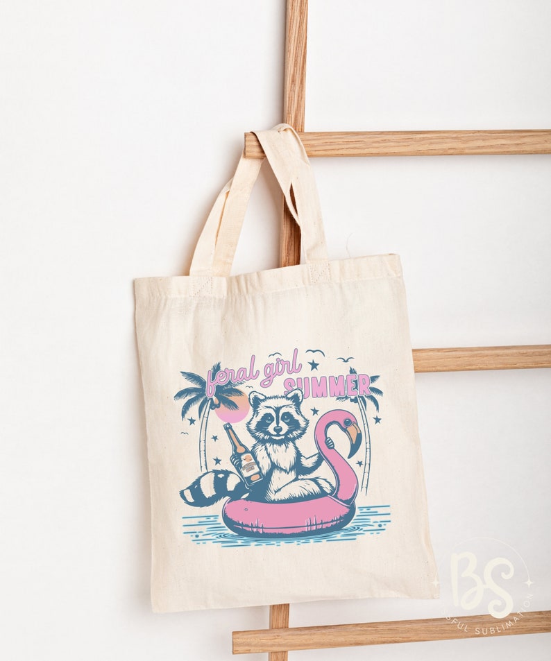 May include: A white tote bag with a pink flamingo pool float and a raccoon holding a bottle of beer. The text "feral girl summer" is printed on the bag.