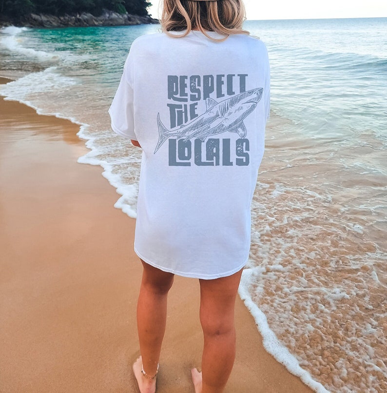 a woman wearing an oversize sweatshirt shirt.  the shirt features a graphic of a shark and the words "respect the locals" in a retro text.  she is standing in front of the ocean