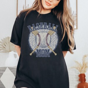 Op de afbeelding: Zwart oversized T-shirt met een grafische print van een honkbal, knuppels en de tekst "Baseball Mom".