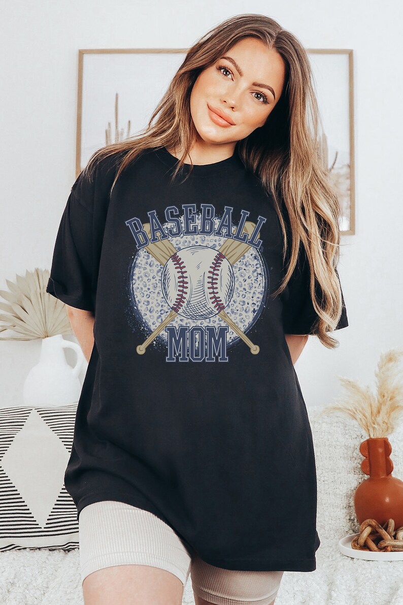 Op de afbeelding: Zwart oversized T-shirt met een grafische print van een honkbal, knuppels en de tekst "Baseball Mom".