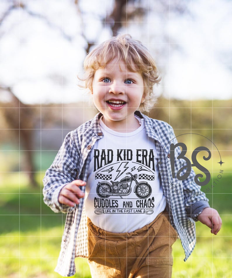 May include: A young boy wearing a white t-shirt with a black and white graphic of a motorcycle and the text "Rad Kid Era Cuddles and Chaos Life in the Fast Lane". He is wearing a plaid shirt over the t-shirt and brown pants. The boy is smiling and looking at the camera.