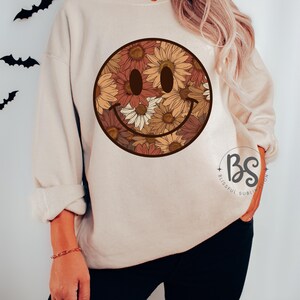 K&ouml;nnte beinhalten: Ein beiger Pullover mit einem floralen Smiley-Gesichtsdesign. Das Smiley-Gesicht besteht aus braunen, orangefarbenen und gelben Blumen.