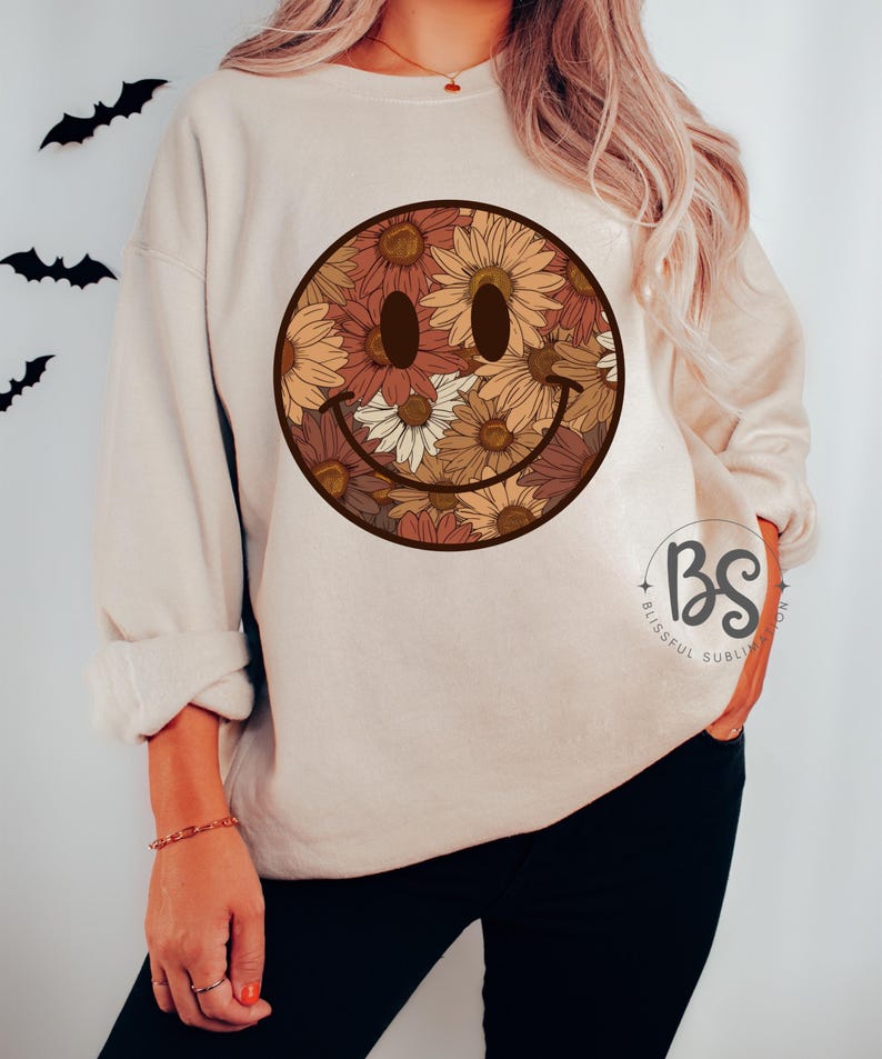 K&ouml;nnte beinhalten: Ein beiger Pullover mit einem floralen Smiley-Gesichtsdesign. Das Smiley-Gesicht besteht aus braunen, orangefarbenen und gelben Blumen.