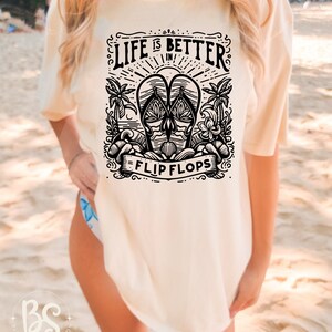 May include: A white t-shirt with a black graphic design that says "Life is better in flip flops". The graphic features a pair of flip flops with palm trees and waves.