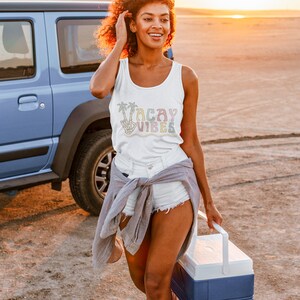 May include: A woman wearing a white tank top with the text "Vacay Vibes" in rainbow colors and palm trees. She is wearing white denim shorts and a gray shirt tied around her waist. She is holding a blue and white cooler.