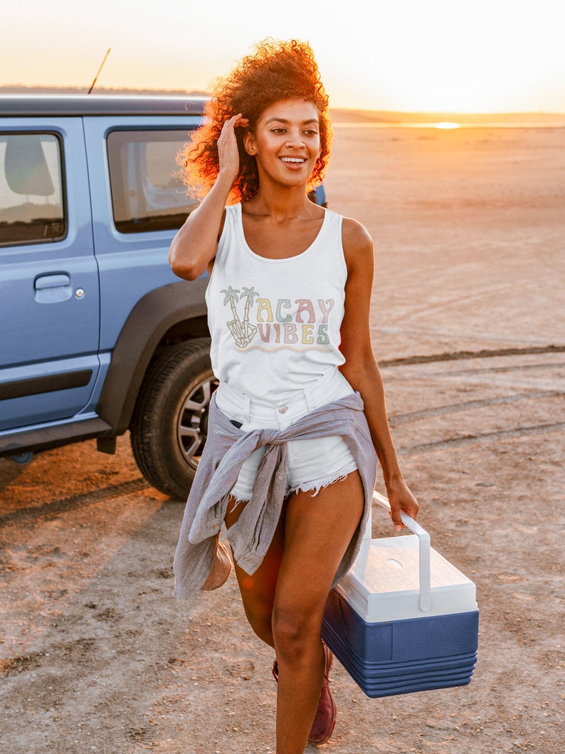 May include: A woman wearing a white tank top with the text "Vacay Vibes" in rainbow colors and palm trees. She is wearing white denim shorts and a gray shirt tied around her waist. She is holding a blue and white cooler.