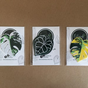 May include: Three iron-on patches featuring different leaf designs. The first patch is white with green and black details. The second patch is black with white details. The third patch is yellow with green and black details. All patches have the text "Botanik" on them.