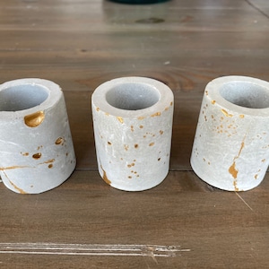 May include: Three small grey concrete candle holders with gold flecks. The holders are cylindrical in shape and have a slightly rough texture.
