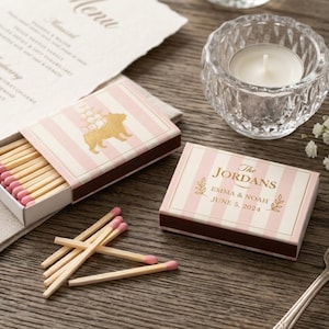 May include: Pink and white striped matchboxes with a gold bear illustration and the text "The Jordans Emma & Noah June 5, 2024". The matches have pink tips. A clear glass candle holder with a white candle is also present.