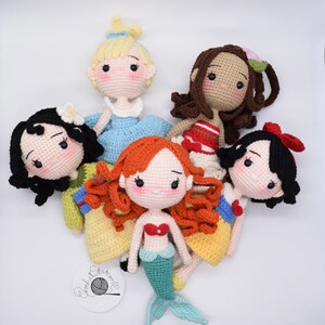 May include: Five crocheted dolls dressed as Disney princesses. The dolls are all wearing different colored dresses and have different hair colors. The dolls are all smiling and have big eyes. The dolls are all sitting on a white background.