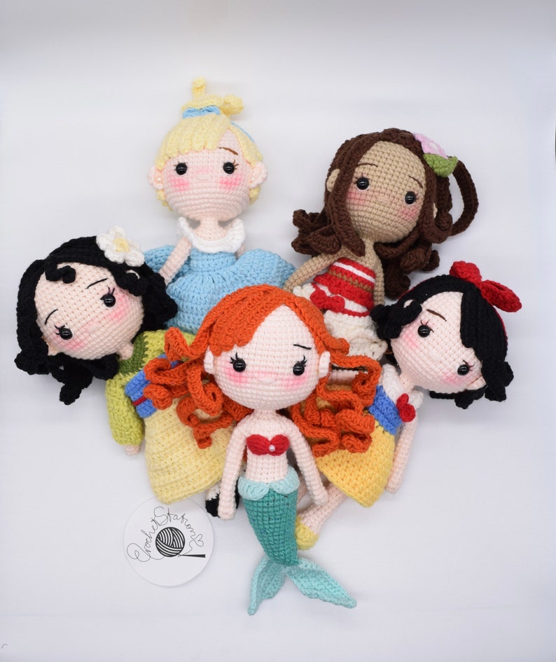 May include: Five crocheted dolls dressed as Disney princesses. The dolls are all wearing different colored dresses and have different hair colors. The dolls are all smiling and have big eyes. The dolls are all sitting on a white background.