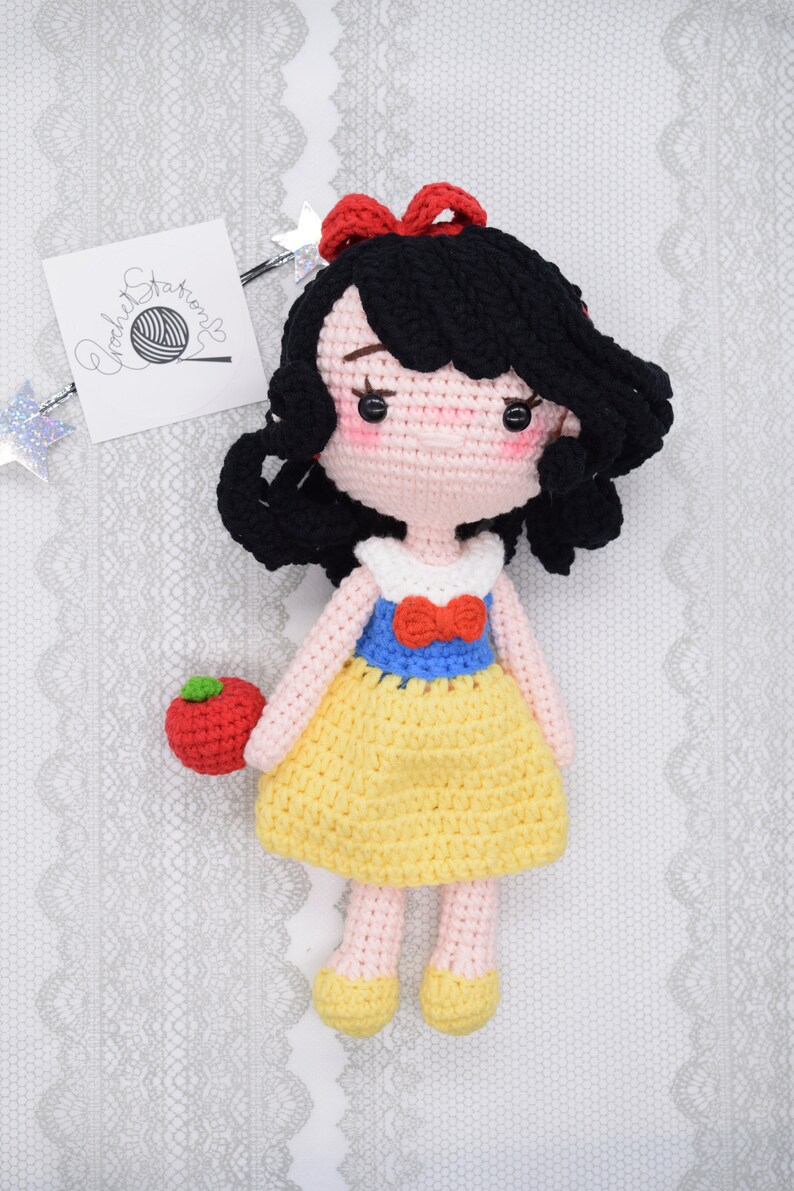 May include: A crocheted Snow White doll with black hair, a red bow, a white and blue dress, and a red apple in her hand.