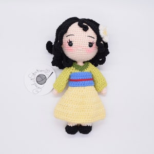 May include: A crocheted doll with black hair, a yellow dress, and a blue and green sash. The doll has a white flower on its head.