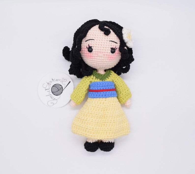 May include: A crocheted doll with black hair, a yellow dress, and a blue and green sash. The doll has a white flower on its head.