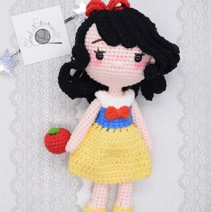 May include: A crocheted Snow White doll with black hair, a red bow, a white and blue dress, and a red apple in her hand.