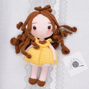May include: A crocheted doll with brown hair, a yellow dress, and brown shoes. The doll is sitting on a white lace background.