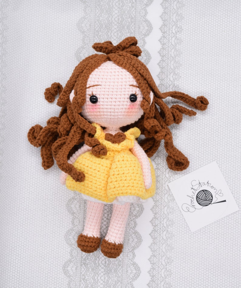May include: A crocheted doll with brown hair, a yellow dress, and brown shoes. The doll is sitting on a white lace background.