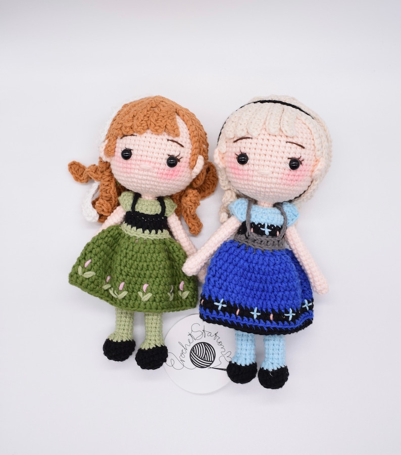 May include: Two crocheted dolls, one with brown hair and a green dress, the other with blonde hair and a blue dress. Both dolls have black eyes and are wearing black shoes.