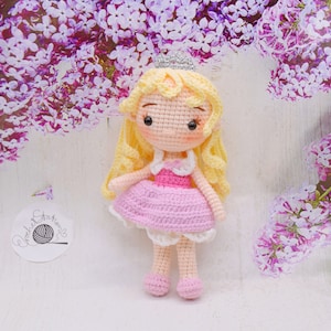 May include: A crocheted doll with blonde hair, a pink dress, and a crown. The doll is standing on a white surface with purple flowers in the background.