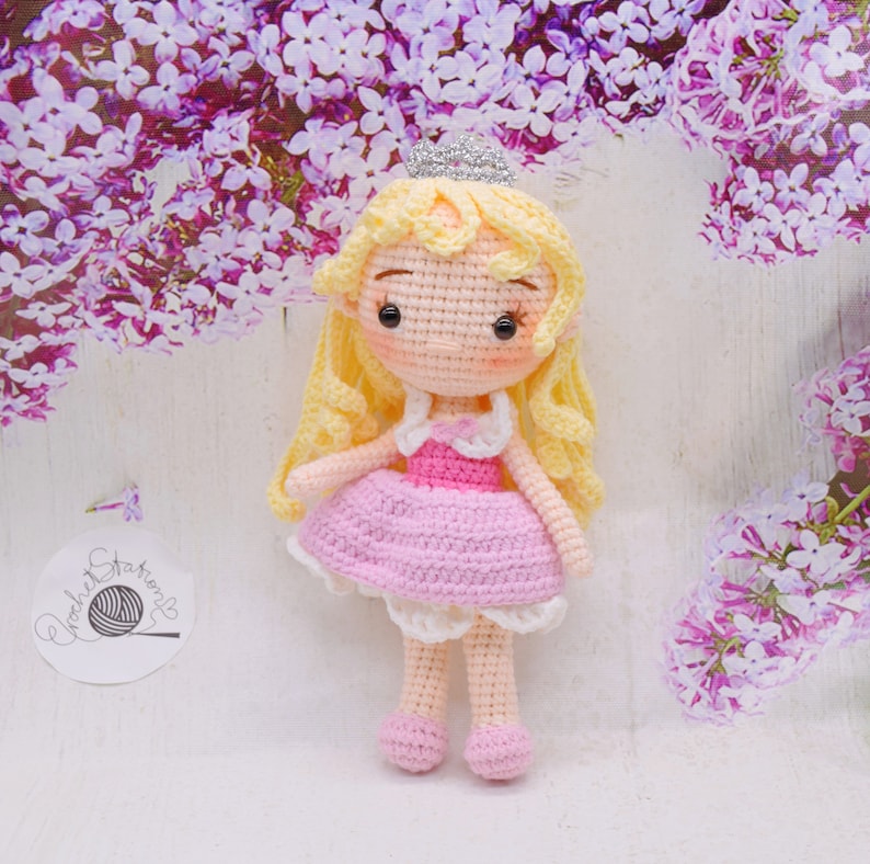 May include: A crocheted doll with blonde hair, a pink dress, and a crown. The doll is standing on a white surface with purple flowers in the background.