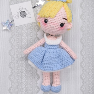 May include: A crocheted doll with blonde hair, a blue dress, and pink shoes. The doll is wearing a blue bow in her hair. The doll is sitting on a white lace background.