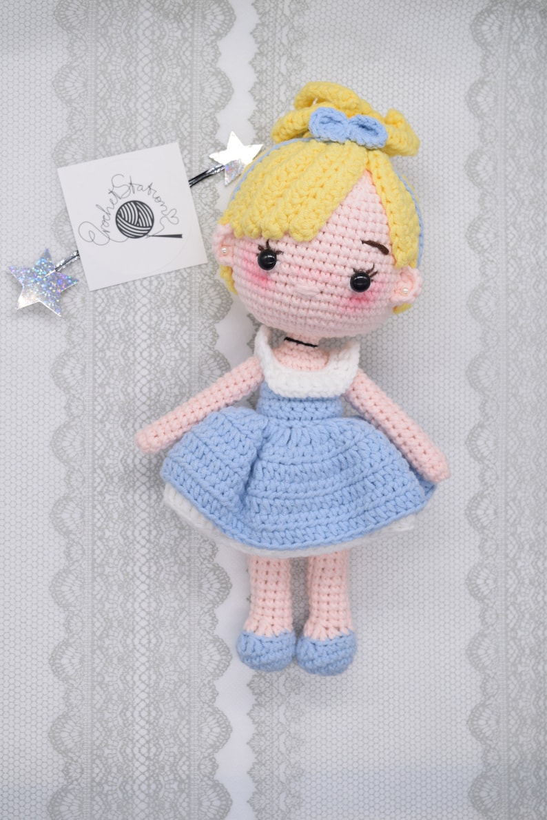 May include: A crocheted doll with blonde hair, a blue dress, and pink shoes. The doll is wearing a blue bow in her hair. The doll is sitting on a white lace background.