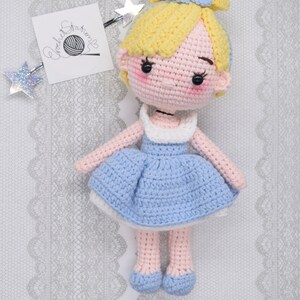 May include: A crocheted doll with blonde hair, a blue dress, and pink shoes. The doll is sitting on a white lace background.