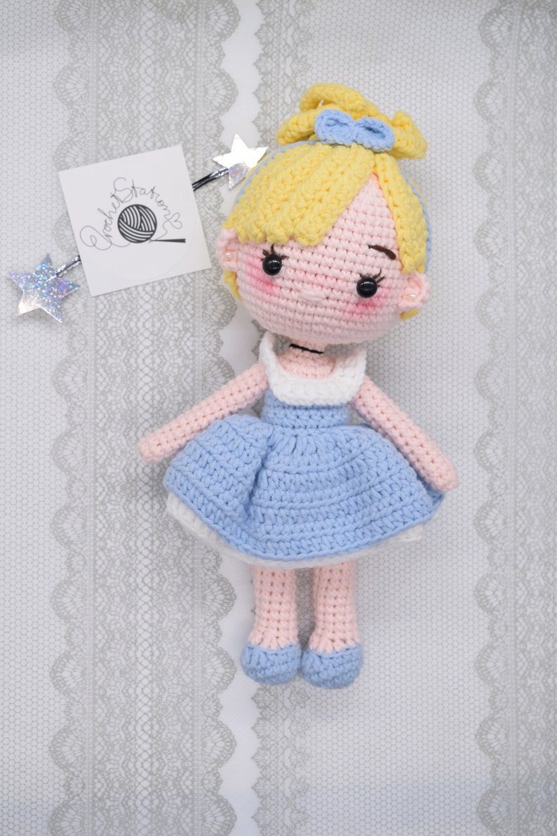 May include: A crocheted doll with blonde hair, a blue dress, and pink shoes. The doll is sitting on a white lace background.